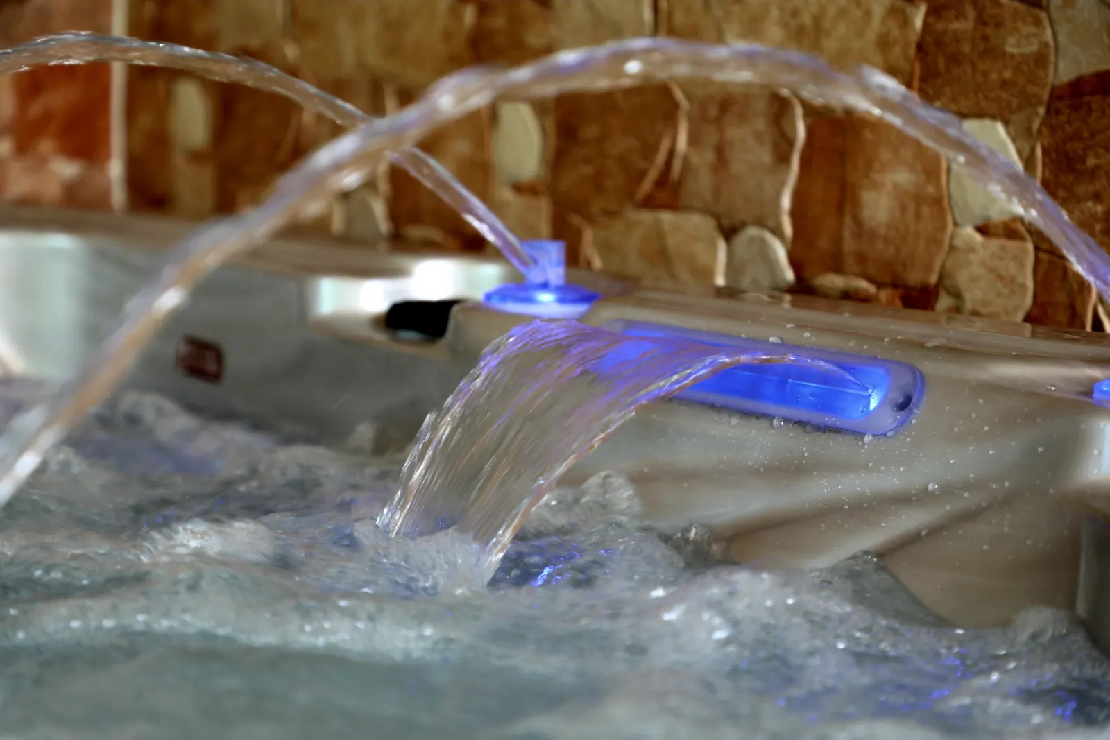 Cadă cu hidromasaj MONAKO, cu un design elegant, având un finisaj lucios, cu jeturi de apă care curg dintr-o zonă iluminată cu LED-uri albastre, situată într-un mediu interior, cu pereți din piatră naturală.