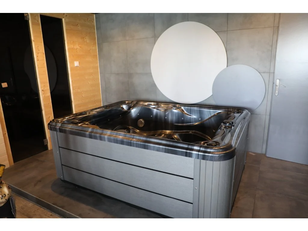 Cadă cu hidromasaj compactă HOME 210×160×77 cm, cu carcasă gri și suprafață acrilică lucioasă, amplasată într-un interior modern, cu pereți de beton și accente de lemn, oferind un spațiu elegant și relaxant pentru 2-3 persoane.