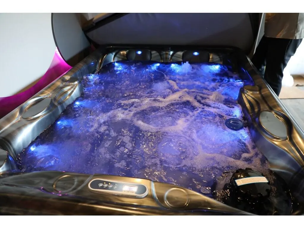 Cadă cu hidromasaj compactă HOME 210×160×77 cm, cu un interior din acril de culoare albastră și lumini LED albastre, situată într-un mediu interior, având margini metalice și un panou de control vizibil.